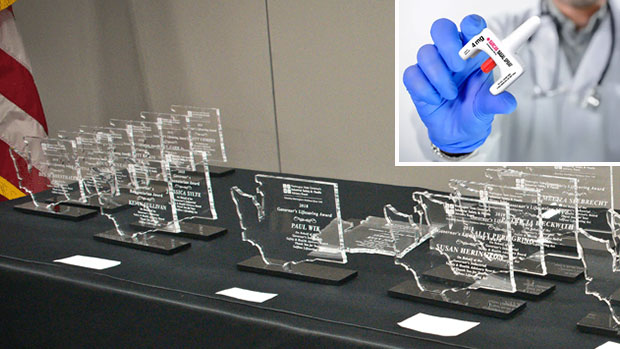 Draped table displaying Governor’s Lifesaving Award statues with inset of doctor holding nalaxone bottle