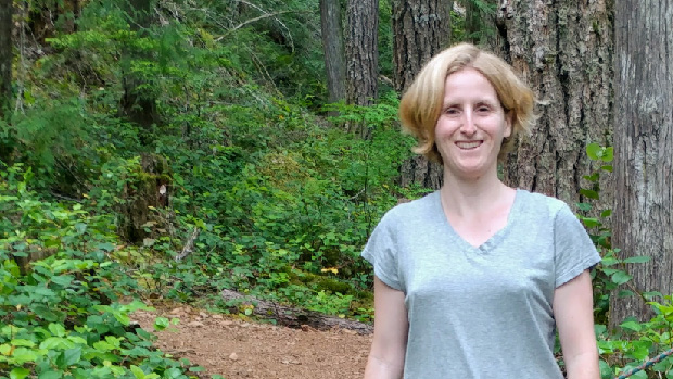 Kelci Mumford, standing in a grove of trees wearing a green t-shirt, smiling