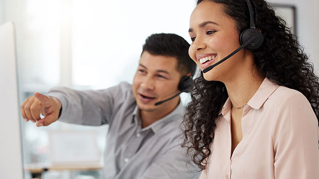 Two people if front of computer. Woman with headset on, smiling. Man, smiling, pointing at the computer.